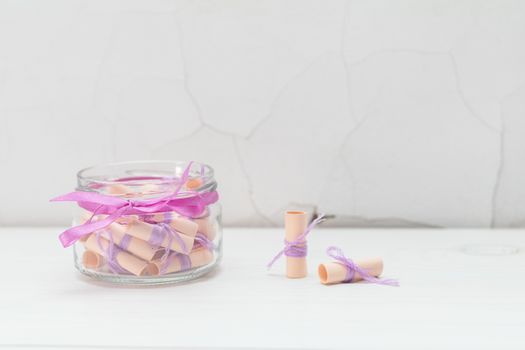 Paper wish scrolls witj violet threads in a glass jar with violet ribbon. White wooden surface close to the white cracked wall. Light tones, copy space.