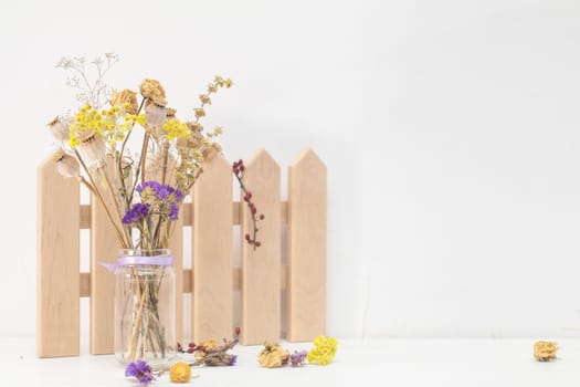 Dried flowers and poppy heads on a stem in a glass jar. Wooden decoration. White wooden table, white cracked wall. Light tones, copy space.