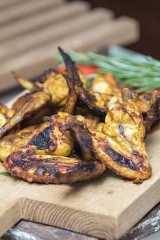 Pile of roasted grilled chicken wings on oak wooden board. Fresh vegetables and greens on the background. Fresh grilled chicken meat. Shallow depth of field.