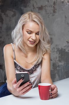 Beautiful blonde girl in home clothes is using a smartphone and smiling while sitting in kitchen