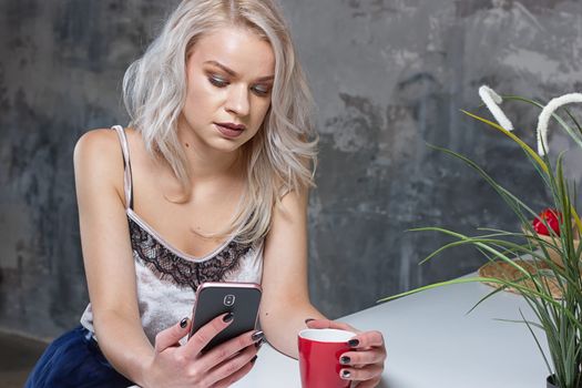 Beautiful blonde girl in home clothes is using a smartphone and smiling while sitting in kitchen