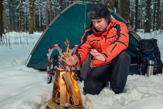 frozen tourist warms his hands near the fire in a tent camp in the winter forest