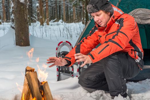 tourist warms his hands near the fire in the winter forest near the tent