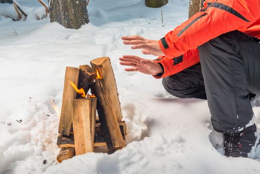 A bonfire in the winter forest and a tourist heating his hands by the fire