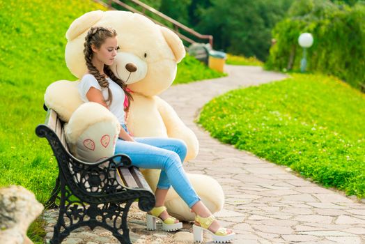 A 20-year-old girl in a park with her huge teddy bear on a bench