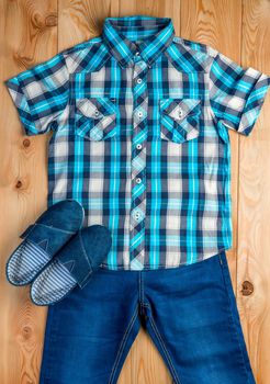 shoes, checkered blue shirt and jeans for a boy set on a wooden floor top view