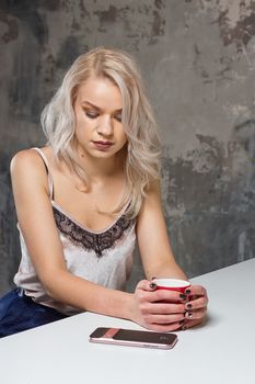 Beautiful blonde girl in home clothes is using a smartphone and smiling while sitting in kitchen