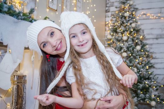 Cute girl in white hat with mother under Christmas tree