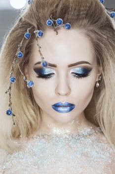 Winter Beauty Woman. Christmas Girl Makeup. Holiday Make-up. Snow Queen High Fashion Portrait over Blue Snow Background. Eyeshadows, False Eyelashes and Crystals on the Lips. Copy Space for Your Text.