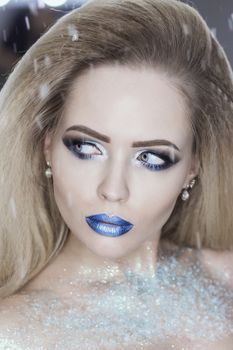 Winter Beauty Woman. Christmas Girl Makeup. Holiday Make-up. Snow Queen High Fashion Portrait over Blue Snow Background. Eyeshadows, False Eyelashes and Crystals on the Lips. Copy Space for Your Text.