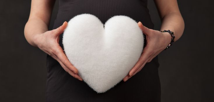 Hands of pregnant woman hold a white heart in front of her abdomen.