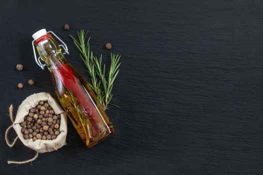 Rosemary bunch, olive oil with pepper and spices and fragrant pepper on black stone surface. Top view, copy space.
