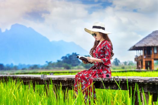 Young woman reading a book and sitting on wooden path with green rice field in Vang Vieng, Laos.