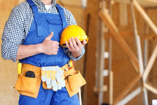 Worker holding hardhat while gesturing thumbs up against workshop