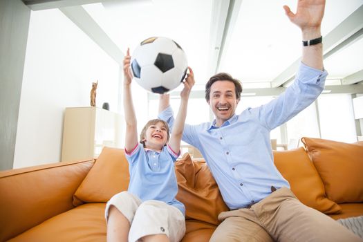 Portrait of happy boy watching soccer match with father on sofa at home