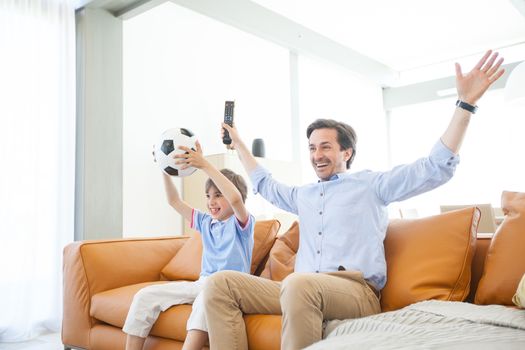 Portrait of happy boy watching soccer match with father on sofa at home