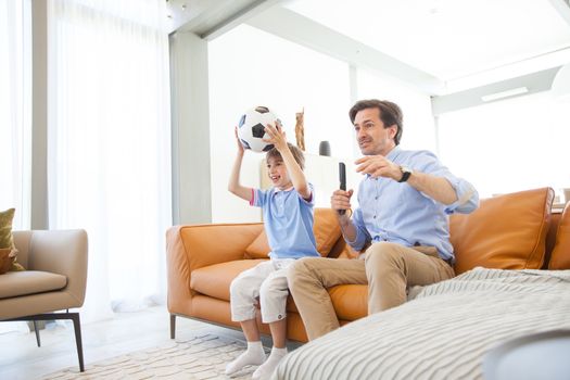 Portrait of happy boy watching soccer match with father on sofa at home