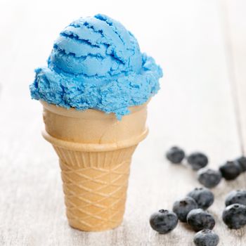 Blue ice cream in waffle cone with blueberry fruit on white rustic wooden background.