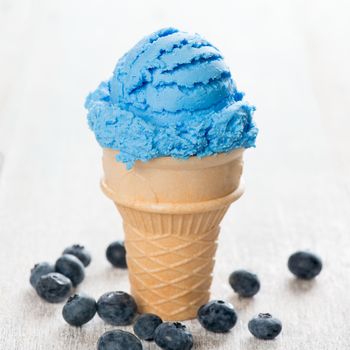 Blue ice cream in waffle cone with blueberry fruit on white wooden background.