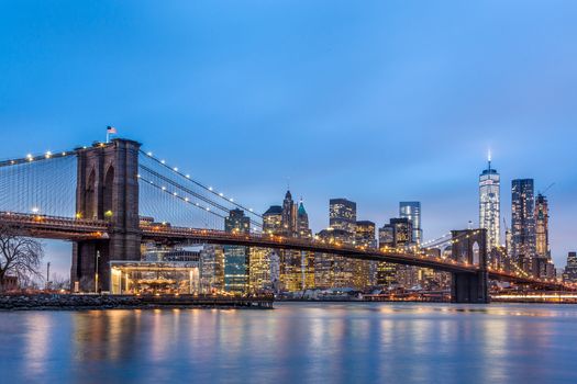 Brooklyn bridge and New York City Manhattan downtown skyline at dusk with skyscrapers illuminated over East River panorama.