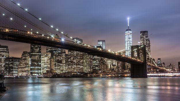 Brooklyn bridge and New York City Manhattan downtown skyline at dusk with skyscrapers illuminated over East River panorama.