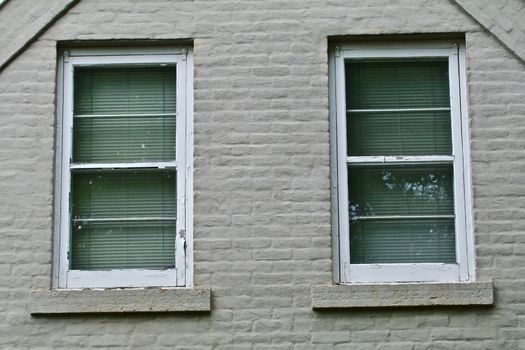 A pair of white windows appear side by side in an old gray painted brick house.