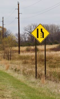 A bright yellow merging lanes warning sign shows against a rural backgound.