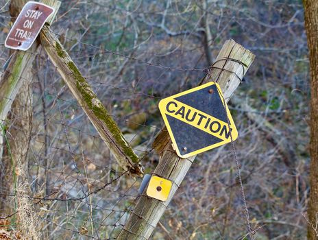 A fence falling into a ravine appears broken and tilted with a stay on trail sign and a bright yellow caution sign.