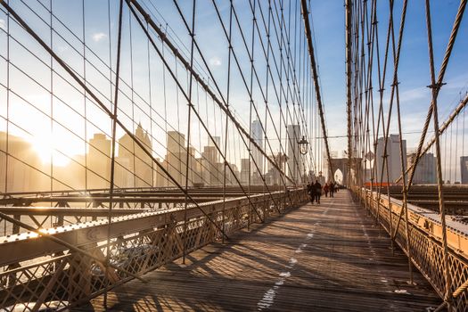 People walking on pedestrian path across Brooklyn bridge. New York City Manhattan downtown skyline in sunset with skyscrapers illuminated over East River panorama as seen from Brooklyn bridge.