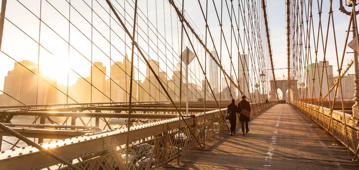 Couple walking on pedestrian path across Brooklyn bridge. New York City Manhattan downtown skyline in sunset with skyscrapers illuminated over East River panorama as seen from Brooklyn bridge.