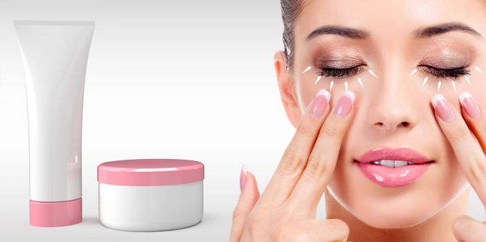 Woman massaging her face with fingers and cosmetics tube and jar on light grey background