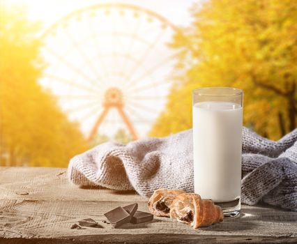 Milk with a croissant on a wooden table. Chunks of chocolate and a knitted sweater next. Blurred background of a ferris wheel in autumn in the park.