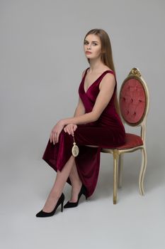 Young beautiful long-haired female model poses in long red dress near red chair on grey background