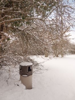 snow covered scene outside winter, public park trees and bin; essex; england; uk