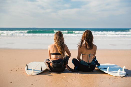 Two beautiful female friends at the beach sitting on the sand with her surfboards and looking to the waves