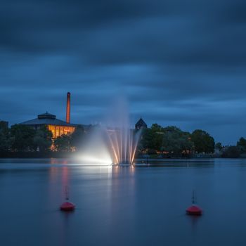 Night landscape with a fountain in the middle of the river. On the back is a former cotton mill.