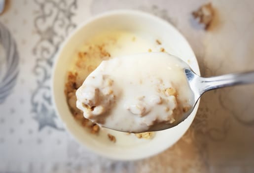 top view of a spoon filled with yogurt and cereal with a bowl and a table set for breakfast in the background