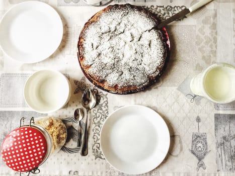 aerial view of a table set for breakfast with white plates, teaspoons and a cake with powdered sugar