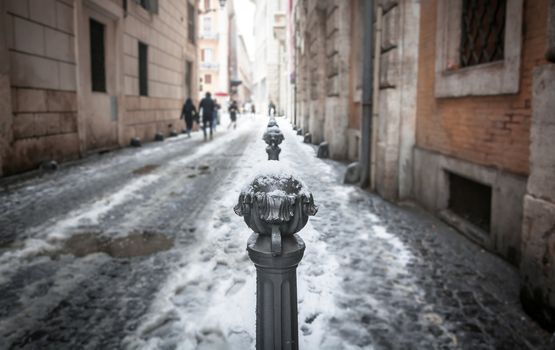 close-up view of a decorated traffic-covered bollard covered in snow in a city alley with people walking in the background