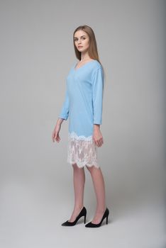 Young beautiful long-haired female model poses in blue dress with white lace on grey background