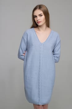 Young beautiful long-haired female model poses in knitted blue dress on grey background