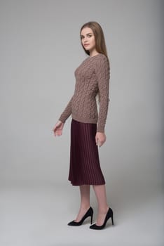 Young beautiful long-haired female model poses in knitted blouse and brown skirt on grey background