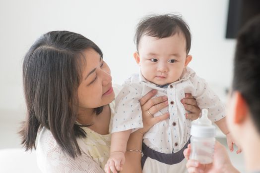 Father showing milk bottle to baby. Happy Asian family at home, candid living lifestyle indoors.
