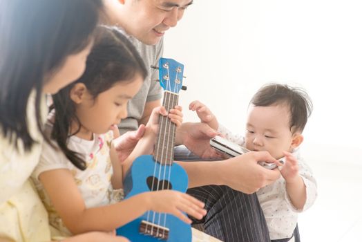 Parents and children singing and playing ukulele and harmonica together. Asian family spending quality time at home, natural living lifestyle indoors.