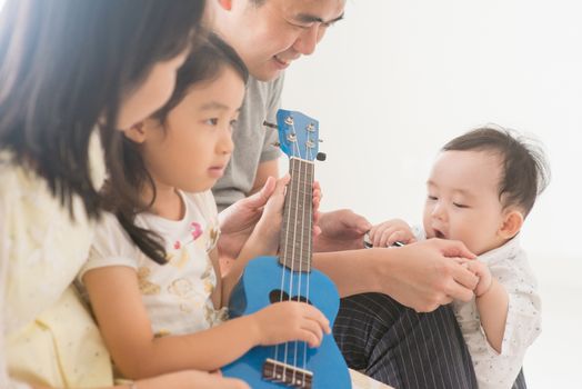 Parents and children playing ukulele together. Asian family spending quality time at home, natural living lifestyle indoors.