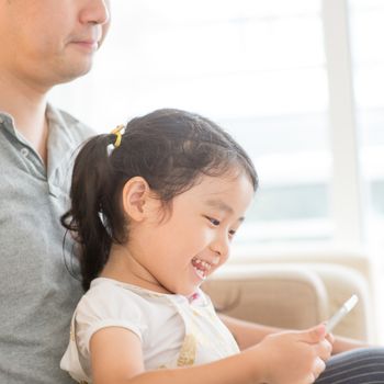 Father using touchscreen tablet PC with daughter on sofa. Asian family at home, living lifestyle indoors.