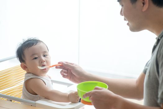 Happy Asian family at home. Father feeding solid food to his 9 months old baby boy in the kitchen, living lifestyle indoors. 