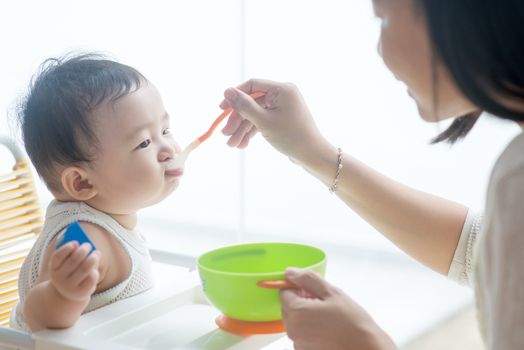 Happy Asian family at home. Mother feeding solid food to 9 months old baby boy in the kitchen, living lifestyle indoors. 