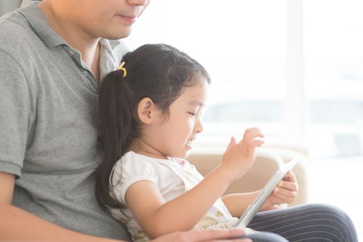 Father and daughter playing with digital tablet on sofa. Asian family at home, living lifestyle indoors.