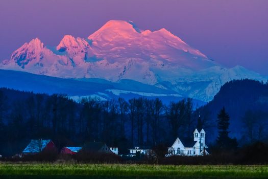 Snow capped Mount Baker at twilight across open field with church in the middle ground